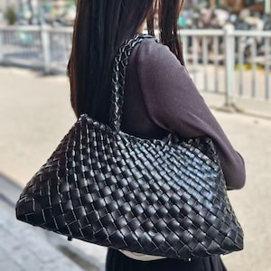 Op de afbeelding: Een zwarte geweven leren tote bag met een gevlochten handvat. De tas heeft een trapeziumvorm en een gestructureerd oppervlak. De tas wordt over de schouder gedragen.