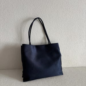 Peut inclure: Un sac fourre-tout bleu marine fonc&eacute; uni de forme rectangulaire avec deux fines anses assorties. Le sac pr&eacute;sente une construction en tissu lisse et mat avec des bords droits et nets, ainsi qu'un panneau avant simple et sans ornement.