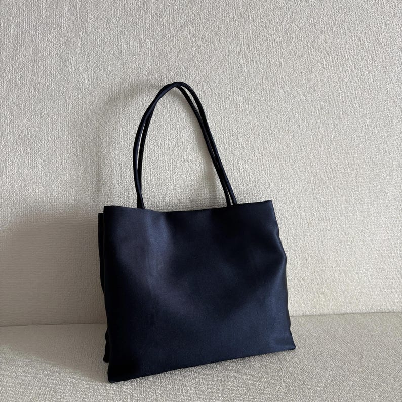 Peut inclure: Un sac fourre-tout bleu marine fonc&eacute; uni de forme rectangulaire avec deux fines anses assorties. Le sac pr&eacute;sente une construction en tissu lisse et mat avec des bords droits et nets, ainsi qu'un panneau avant simple et sans ornement.