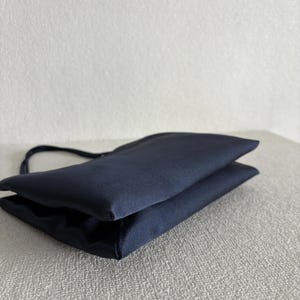 Peut inclure: Un sac &agrave; main bleu marine fonc&eacute; avec une finition lisse et satin&eacute;e. Le sac pr&eacute;sente une silhouette rectangulaire pli&eacute;e aux lignes &eacute;pur&eacute;es et une fine anse unique fix&eacute;e sur le c&ocirc;t&eacute;, mettant en valeur une texture de tissu uniforme et &eacute;l&eacute;gante.