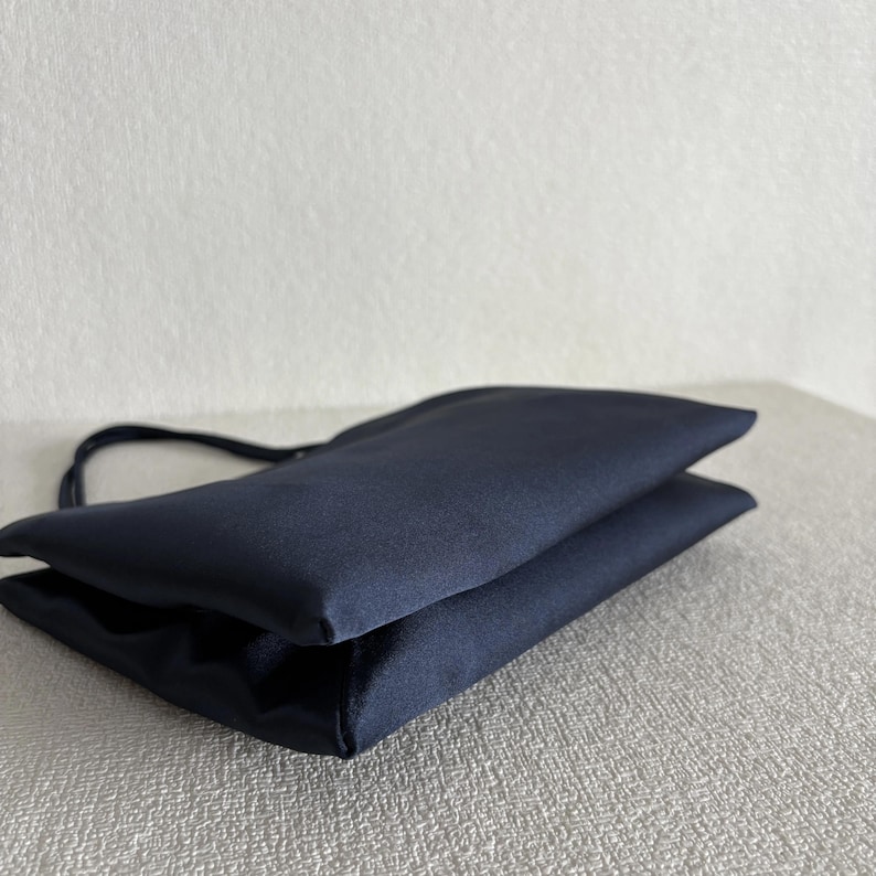 Peut inclure: Un sac &agrave; main bleu marine fonc&eacute; avec une finition lisse et satin&eacute;e. Le sac pr&eacute;sente une silhouette rectangulaire pli&eacute;e aux lignes &eacute;pur&eacute;es et une fine anse unique fix&eacute;e sur le c&ocirc;t&eacute;, mettant en valeur une texture de tissu uniforme et &eacute;l&eacute;gante.