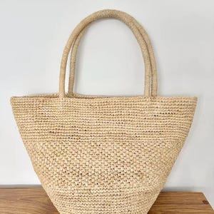 May include: A natural-toned woven tote bag with two sturdy handles. The bag has a wide, open top and a rounded bottom. The woven material appears to be made of straw or a similar natural fiber, creating a casual, beachy aesthetic.