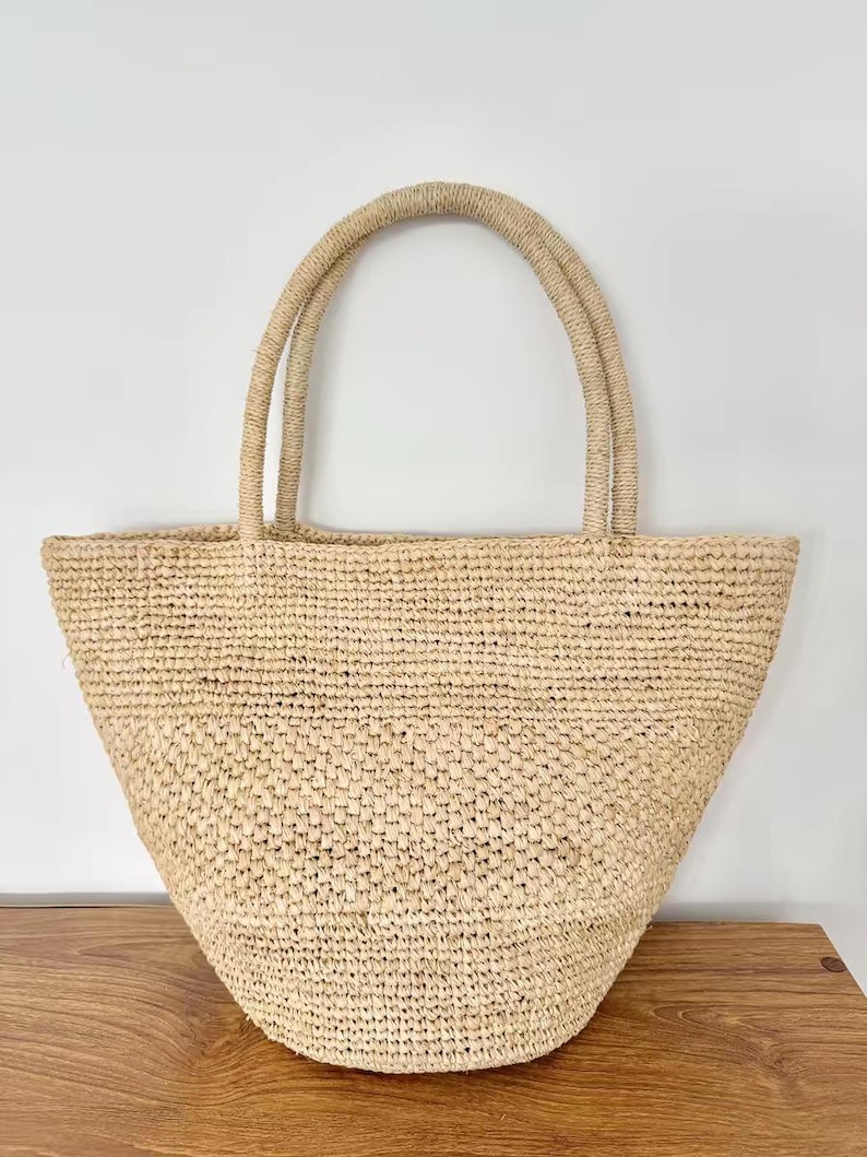 May include: A natural-toned woven tote bag with two sturdy handles. The bag has a wide, open top and a rounded bottom. The woven material appears to be made of straw or a similar natural fiber, creating a casual, beachy aesthetic.