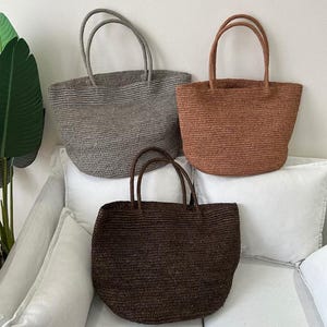 May include: Three woven tote bags in shades of gray, brown, and dark brown. Each bag has two handles and a rounded shape. The bags are made of a textured material and are displayed on a white surface.