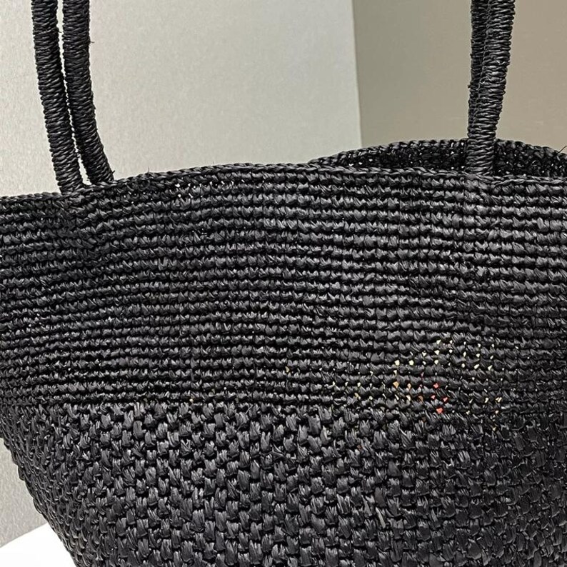 May include: A close-up of a black woven tote bag with two sturdy handles. The bag is made of a tightly woven material, creating a textured surface. The handles are also woven, matching the bag's overall design. The bag is designed for carrying items.