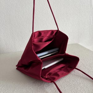 Peut inclure: Un sac &agrave; main en satin bordeaux profond avec une silhouette souple et non structur&eacute;e et de fines bandouli&egrave;res assorties. Le dessus ouvert r&eacute;v&egrave;le un compartiment int&eacute;rieur spacieux doubl&eacute; du m&ecirc;me tissu, contenant deux objets blancs fins.