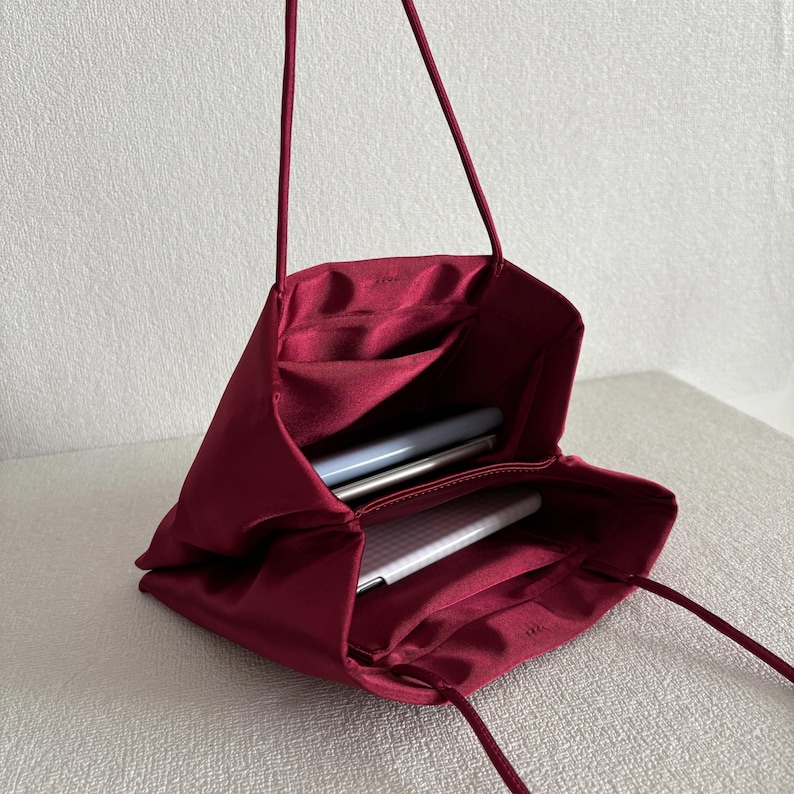 Peut inclure: Un sac &agrave; main en satin bordeaux profond avec une silhouette souple et non structur&eacute;e et de fines bandouli&egrave;res assorties. Le dessus ouvert r&eacute;v&egrave;le un compartiment int&eacute;rieur spacieux doubl&eacute; du m&ecirc;me tissu, contenant deux objets blancs fins.