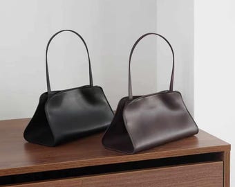 Puede incluir: Dos bolsos estructurados con acabado liso, uno en negro y otro en marrón oscuro. Cada bolso presenta una base ancha y abierta, una abertura superior curva y una sola asa fina y arqueada fijada en el centro, creando una silueta elegante.
