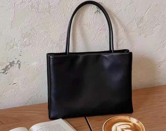 May include: A black rectangular handbag with a smooth finish and two thin, rounded top handles. The bag features a structured, minimalist silhouette with clean lines and a solid black exterior, designed for carrying personal items in a simple style.