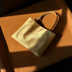 Peut inclure: Un petit sac &agrave; main rectangulaire confectionn&eacute; dans un tissu satin&eacute; dor&eacute; p&acirc;le et brillant. Le sac pr&eacute;sente une silhouette simple et structur&eacute;e avec deux fines anses assorties de couleur dor&eacute;e fix&eacute;es sur le dessus, offrant un aspect &eacute;pur&eacute;.