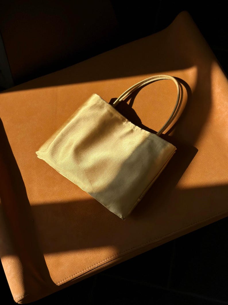 Peut inclure: Un petit sac &agrave; main rectangulaire confectionn&eacute; dans un tissu satin&eacute; dor&eacute; p&acirc;le et brillant. Le sac pr&eacute;sente une silhouette simple et structur&eacute;e avec deux fines anses assorties de couleur dor&eacute;e fix&eacute;es sur le dessus, offrant un aspect &eacute;pur&eacute;.