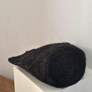 May include: A black woven tote bag with a cylindrical shape and a flat, slightly angled top. The bag is made of a tightly woven material, and the bottom features a spiral pattern. The bag is sitting on a white surface.