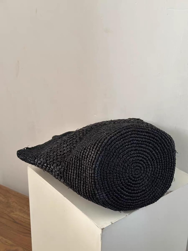 May include: A black woven tote bag with a cylindrical shape and a flat, slightly angled top. The bag is made of a tightly woven material, and the bottom features a spiral pattern. The bag is sitting on a white surface.