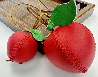 Peut inclure: Deux ornements en forme de pomme en cuir rouge avec des feuilles en cuir vert et des tiges en cuir marron. Les pommes ont un design de couture de type baseball. Une pomme est plus grande que l'autre. Les ornements sont sur un plateau en bois brun.