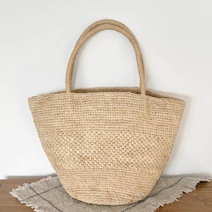 May include: A woven straw tote bag with two rounded handles. The bag is a light tan color and sits on a neutral-colored, fringed cloth. The bag's texture is visible, and the handles are made of a twisted rope-like material.