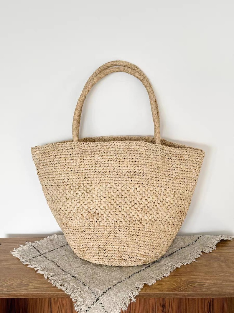 May include: A woven straw tote bag with two rounded handles. The bag is a light tan color and sits on a neutral-colored, fringed cloth. The bag's texture is visible, and the handles are made of a twisted rope-like material.