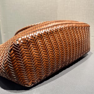 以下が含まれることがあります： 長方形のベースと編み込みのハンドルが付いた、茶色の織りトートバッグ。バッグの複雑な織り模様が、テクスチャード加工された表面を作り出しています。バッグは天然素材で作られています。
