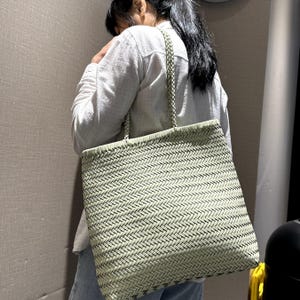 Può includere: Una borsa tote intrecciata in un motivo verde chiaro e nero. La borsa ha una forma rettangolare e un manico intrecciato. La borsa viene portata a spalla.