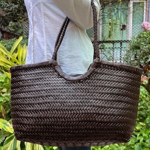 Puede incluir: Un bolso tote de color marrón oscuro con un patrón de espiga trenzado en todo el exterior. El bolso tiene dos correas trenzadas para el hombro unidas al borde superior, que se curva hacia abajo en el centro para formar la abertura.