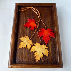 May include: A wooden tray with a dark brown finish, featuring three maple leaf ornaments in shades of red and yellow. The leaves are attached to a thin, brown leather cord. The tray has a rectangular shape with a raised border.