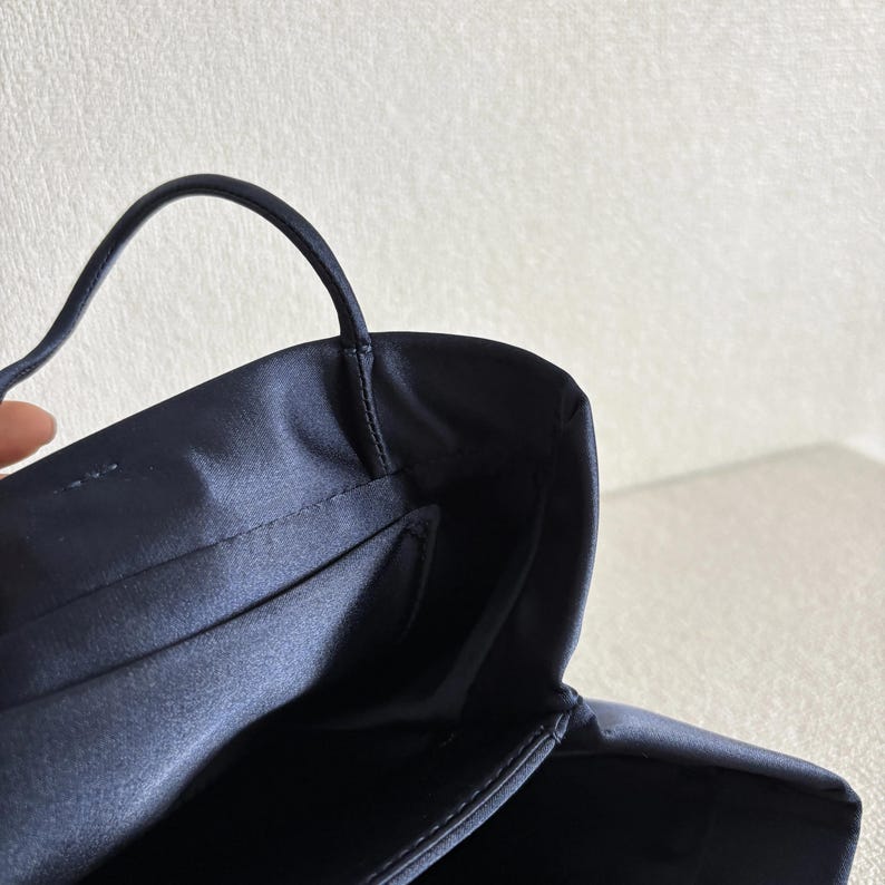 Peut inclure: Un sac &agrave; main en satin bleu marine fonc&eacute; dot&eacute; d'une finition lisse et lustr&eacute;e, d'une fine poign&eacute;e sup&eacute;rieure unique et d'une ouverture structur&eacute;e. L'int&eacute;rieur est doubl&eacute; d'un tissu bleu marine assorti avec des coutures nettes sur les bords.