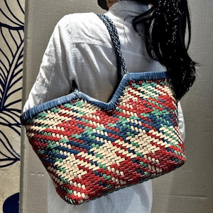 May include: A colourful woven tote bag with a pattern of red, blue, green, and cream. The bag has blue leather trim and braided straps. The bag is carried over the shoulder.