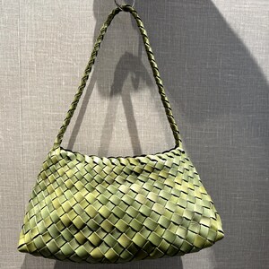 Peut inclure: Un sac à main vert tissé avec une poignée tressée. Le sac est fait de bandes entrelacées, créant un aspect texturé en forme de panier. Le sac est de couleur vert clair.