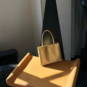 Peut inclure: Un petit sac &agrave; main rectangulaire de couleur jaune dor&eacute; avec une finition lisse et satin&eacute;e. Le sac est dot&eacute; de deux fines anses assorties fix&eacute;es sur le bord sup&eacute;rieur, offrant une silhouette simple et structur&eacute;e aux lignes &eacute;pur&eacute;es.