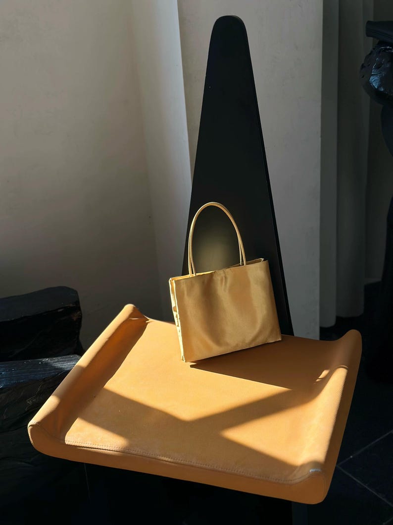 Peut inclure: Un petit sac &agrave; main rectangulaire de couleur jaune dor&eacute; avec une finition lisse et satin&eacute;e. Le sac est dot&eacute; de deux fines anses assorties fix&eacute;es sur le bord sup&eacute;rieur, offrant une silhouette simple et structur&eacute;e aux lignes &eacute;pur&eacute;es.