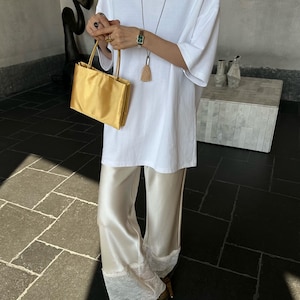 Peut inclure: Un t-shirt blanc &agrave; manches courtes associ&eacute; &agrave; un pantalon large en satin cr&egrave;me avec une bordure en dentelle blanche aux ourlets. La tenue comprend un petit sac &agrave; main dor&eacute;, un long collier avec un pendentif &agrave; pompon et des chaussures dor&eacute;es.