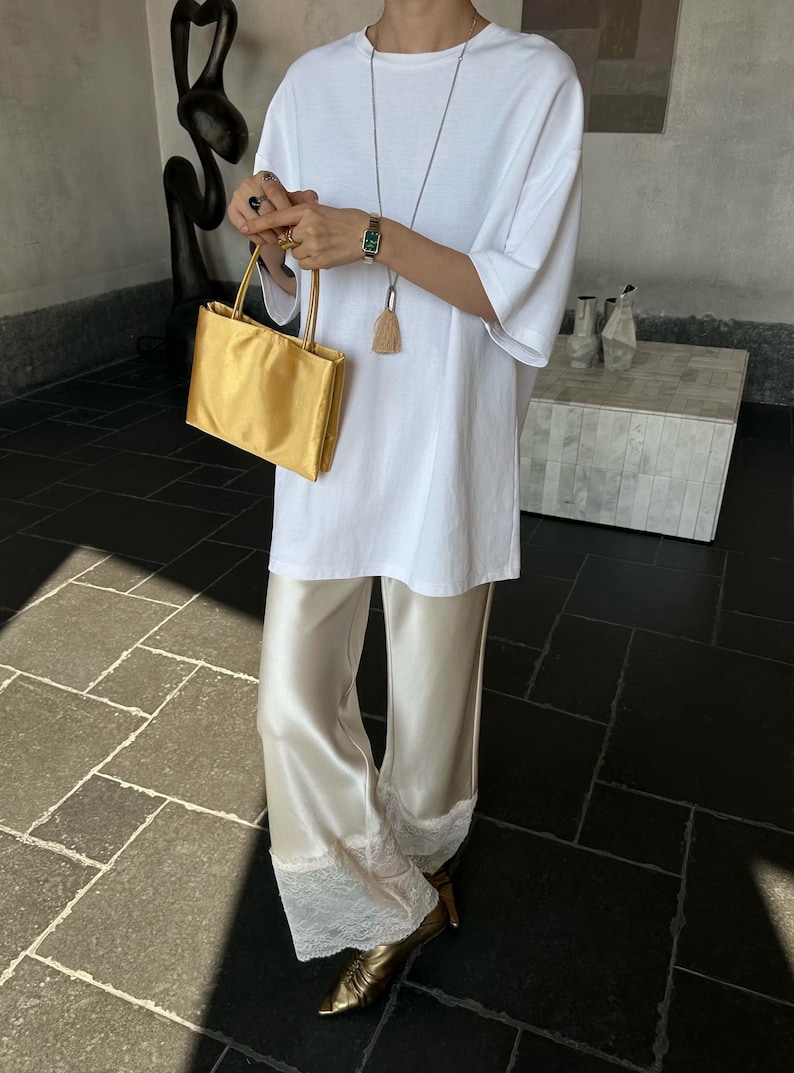 Peut inclure: Un t-shirt blanc &agrave; manches courtes associ&eacute; &agrave; un pantalon large en satin cr&egrave;me avec une bordure en dentelle blanche aux ourlets. La tenue comprend un petit sac &agrave; main dor&eacute;, un long collier avec un pendentif &agrave; pompon et des chaussures dor&eacute;es.