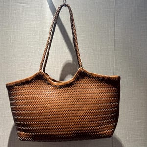 以下が含まれることがあります： 茶色の編み込みトートバッグ、編み込みハンドル付き。 バッグは長方形で、上端が湾曲しています。 編み込み模様がテクスチャード加工された表面を作り出しています。 バッグは金属製のフックから吊り下げられています。