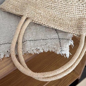 May include: A woven straw tote bag with two rounded handles. The bag is a light beige color and appears to be made of natural fibers. The handles are also made of woven straw. The bag is sitting on a light gray fabric.