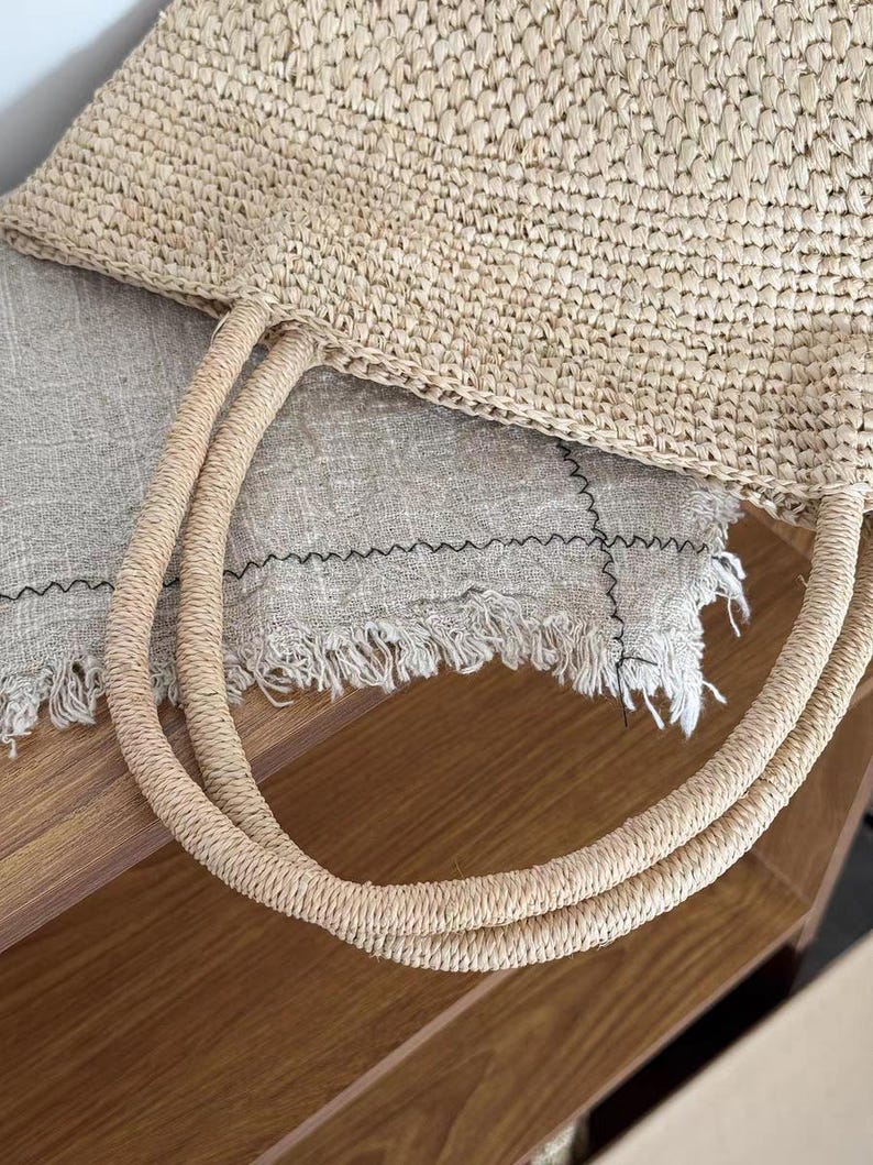 May include: A woven straw tote bag with two rounded handles. The bag is a light beige color and appears to be made of natural fibers. The handles are also made of woven straw. The bag is sitting on a light gray fabric.
