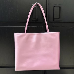Peut inclure: Un sac fourre-tout rectangulaire d'une couleur rose clair unie avec une finition lisse et satin&eacute;e. Le sac pr&eacute;sente deux fines bretelles assorties de couleur rose fix&eacute;es au bord sup&eacute;rieur, offrant un design simple et &eacute;pur&eacute; pour transporter des objets.