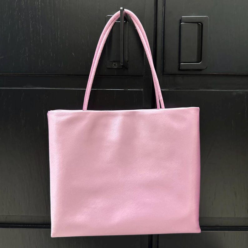 Peut inclure: Un sac fourre-tout rectangulaire d'une couleur rose clair unie avec une finition lisse et satin&eacute;e. Le sac pr&eacute;sente deux fines bretelles assorties de couleur rose fix&eacute;es au bord sup&eacute;rieur, offrant un design simple et &eacute;pur&eacute; pour transporter des objets.