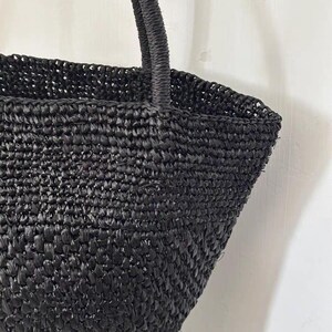 May include: A black woven tote bag with a structured shape and a sturdy handle. The bag is made of a tightly woven material, creating a textured surface. The handle is also woven, providing a cohesive design. The bag is suitable for carrying various items.