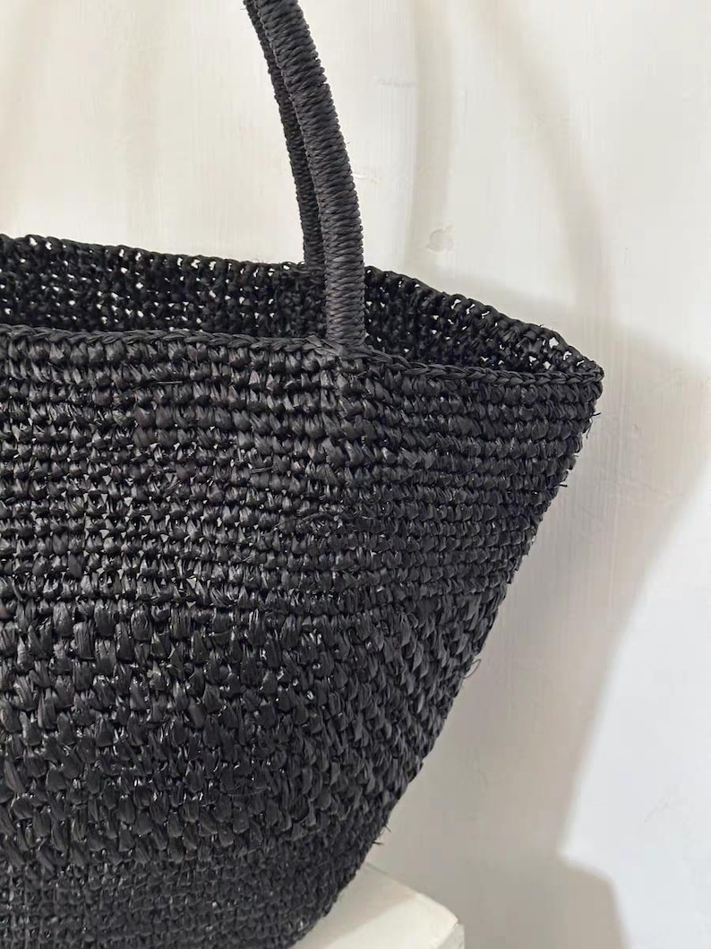 May include: A black woven tote bag with a structured shape and a sturdy handle. The bag is made of a tightly woven material, creating a textured surface. The handle is also woven, providing a cohesive design. The bag is suitable for carrying various items.