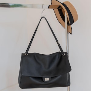 May include: A black leather shoulder bag with a pebbled texture, a fold-over flap, and a silver-toned rectangular turn-lock closure. The bag features a single adjustable shoulder strap attached with silver metal rings and hardware.