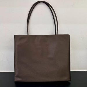 Peut inclure: Un sac cabas marron fonc&eacute; uni avec un corps rectangulaire et deux fines anses assorties. Le sac pr&eacute;sente une finition lisse et mate ainsi qu'un design &eacute;pur&eacute; et structur&eacute; avec une base plate et des bords droits, offrant un aspect simple et fonctionnel.
