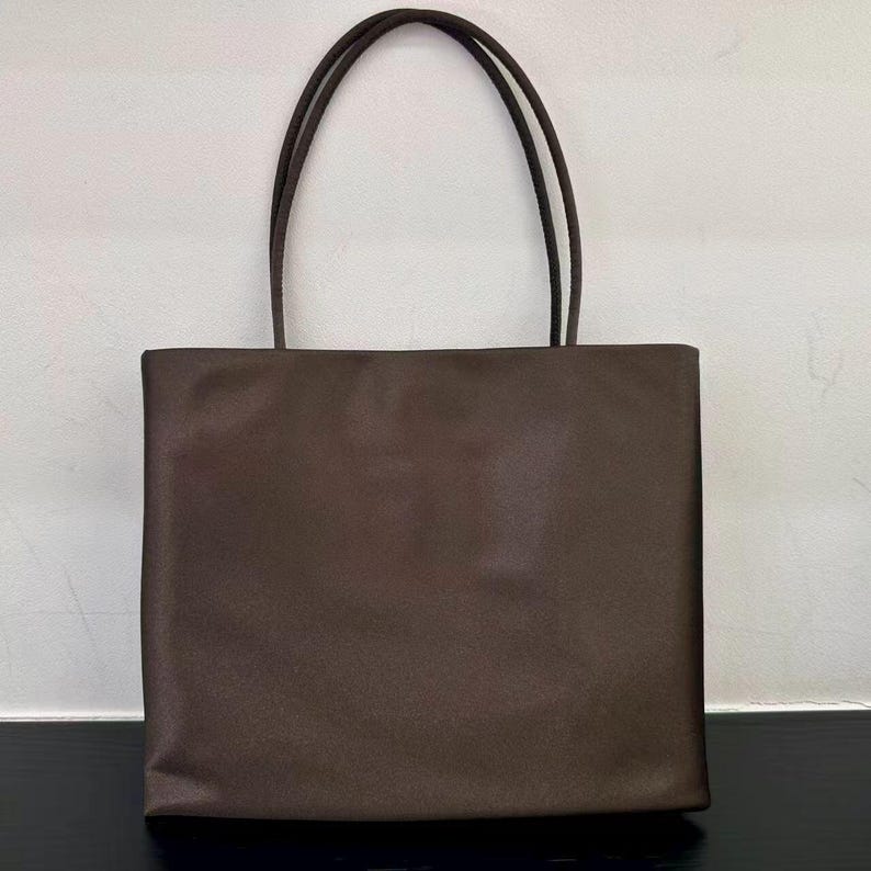 Peut inclure: Un sac cabas marron fonc&eacute; uni avec un corps rectangulaire et deux fines anses assorties. Le sac pr&eacute;sente une finition lisse et mate ainsi qu'un design &eacute;pur&eacute; et structur&eacute; avec une base plate et des bords droits, offrant un aspect simple et fonctionnel.