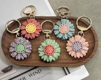Peut inclure: Six porte-clés en forme de tournesol colorés avec des attaches dorées sont disposés sur un plateau en bois. Chaque porte-clés présente une combinaison de couleurs différente, dont rose, violet, vert et bleu, avec un centre contrasté.