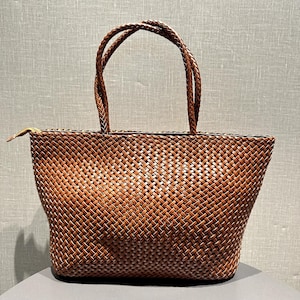 Puede incluir: Un bolso tote tejido marrón con asas trenzadas. El bolso tiene forma rectangular con cierre de cremallera. El patrón de tejido es intrincado, creando una superficie texturizada. Las asas también están trenzadas.