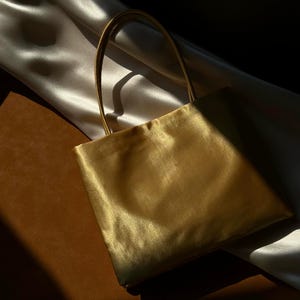 Peut inclure: Un sac &agrave; main de forme carr&eacute;e fabriqu&eacute; dans un tissu dor&eacute; m&eacute;tallique et lustr&eacute;. Le sac pr&eacute;sente deux fines anses assorties fix&eacute;es sur le bord sup&eacute;rieur. Le mat&eacute;riau poss&egrave;de une surface lisse et r&eacute;fl&eacute;chissante qui capte la lumi&egrave;re.
