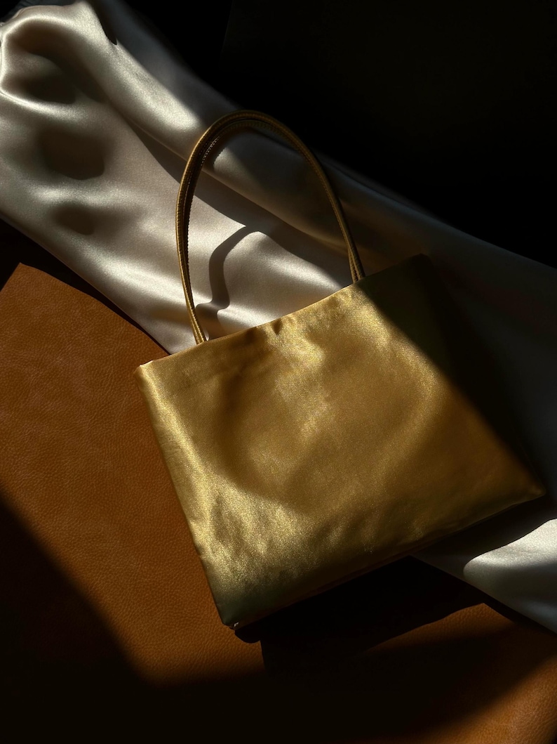 Peut inclure: Un sac &agrave; main de forme carr&eacute;e fabriqu&eacute; dans un tissu dor&eacute; m&eacute;tallique et lustr&eacute;. Le sac pr&eacute;sente deux fines anses assorties fix&eacute;es sur le bord sup&eacute;rieur. Le mat&eacute;riau poss&egrave;de une surface lisse et r&eacute;fl&eacute;chissante qui capte la lumi&egrave;re.