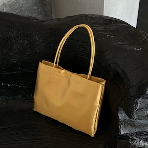 Puede incluir: Un bolso de mano rectangular con acabado en oro metálico y una textura suave similar al satén. El bolso cuenta con dos asas finas y redondeadas unidas al borde superior, ofreciendo una apariencia estructurada y suave con líneas limpias.