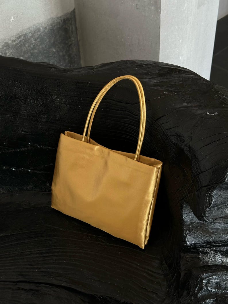 Peut inclure: Un sac &agrave; main rectangulaire au fini dor&eacute; m&eacute;tallique avec une texture lisse rappelant le satin. Le sac pr&eacute;sente deux anses fines et arrondies fix&eacute;es au bord sup&eacute;rieur, offrant une apparence structur&eacute;e et douce avec des lignes &eacute;pur&eacute;es.