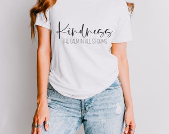 May include: White t-shirt featuring the word "Kindness" in a cursive script, with the text "THE CALM IN ALL STORMS" beneath. The shirt is paired with blue jeans.