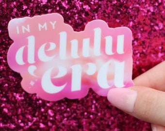 May include: A pink and white sticker with the text "In my delulu era" on a pink glitter background.