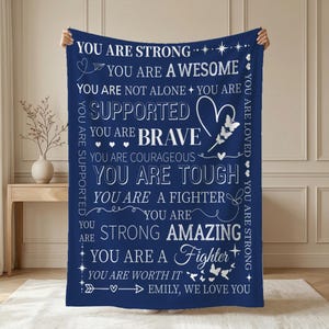 Könnte beinhalten: Eine marineblaue Decke mit weißem Text, der ermutigende Sätze wie "You Are Strong" und "You Are Brave" enthält. Das Design zeigt ein Herz und Schmetterlinge, mit der Botschaft "Emily, We Love You" am unteren Rand.